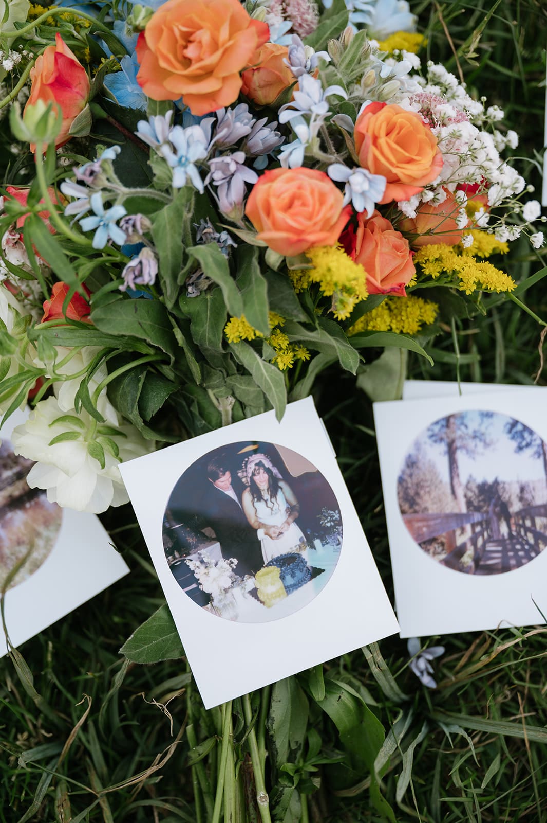 A bouquet of orange and blue flowers lies on grass with three instant photos placed in front of it.