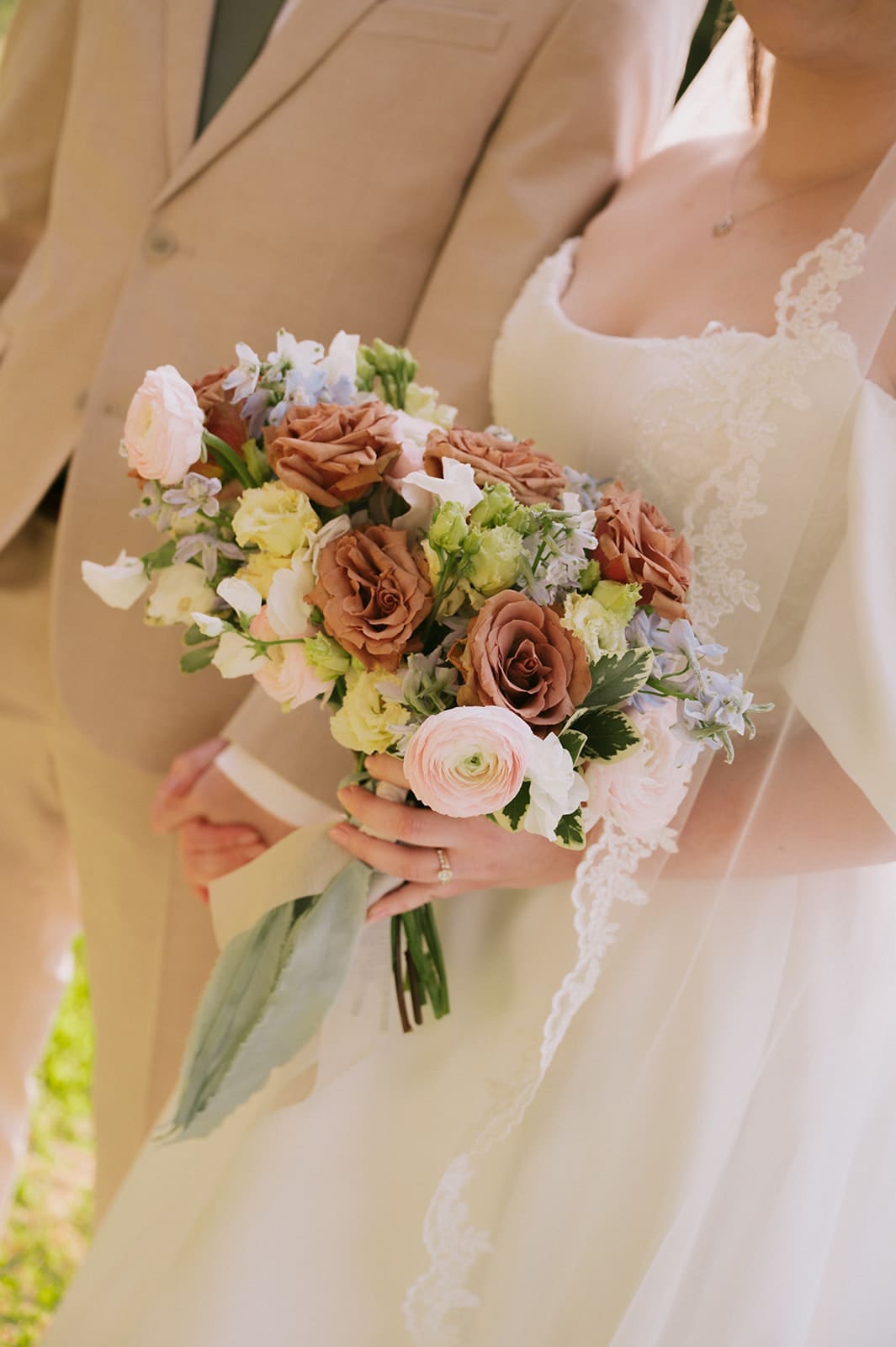A bride in a white dress holds a bouquet of pastel and brown flowers, standing next to a person in a beige suit.