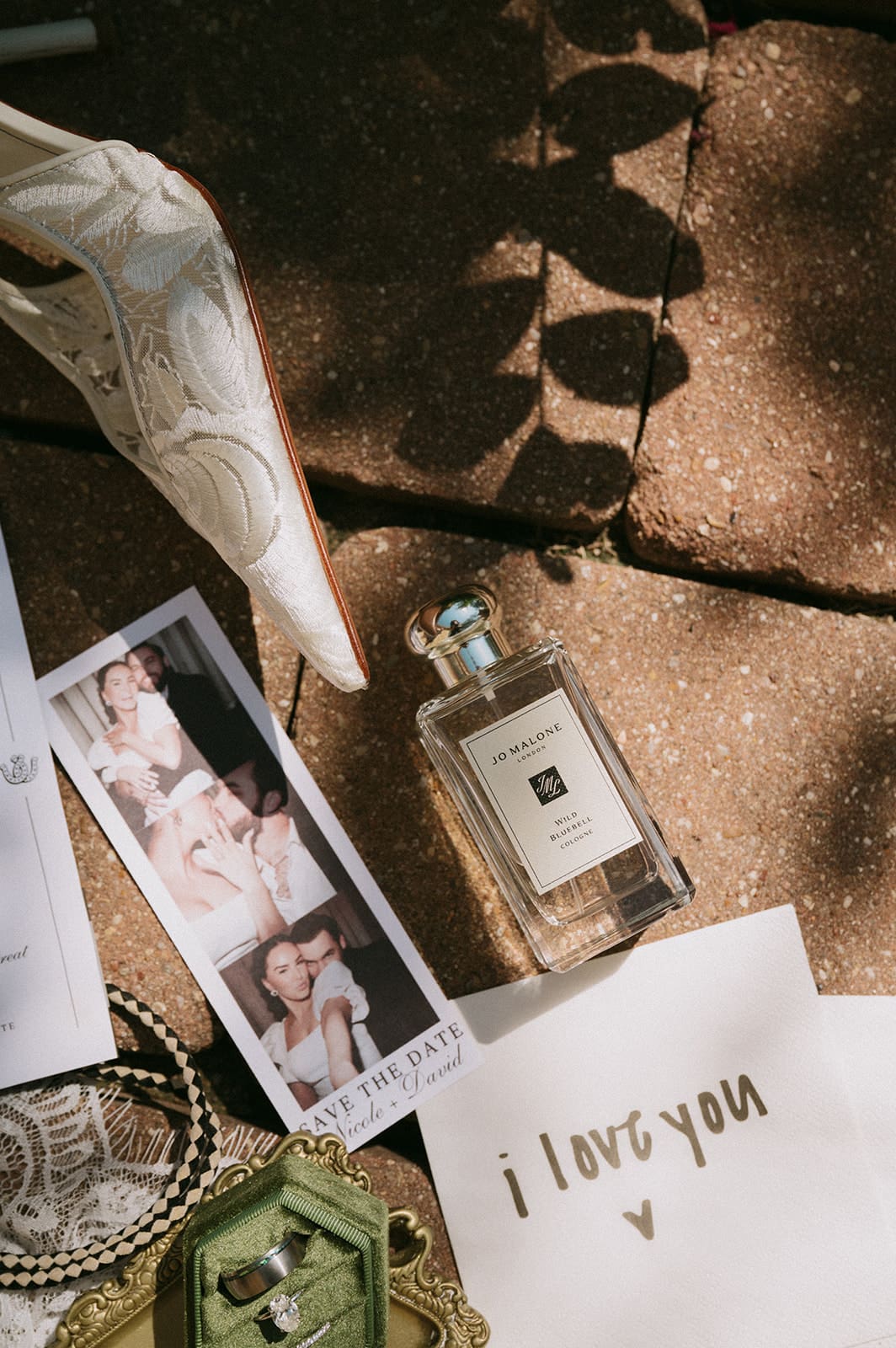 A flat lay of invitation cards, a green notebook, a small box, perfume bottles, a lace cloth, a ribboned pouch, and a photo on a stone surface with some foliage shadows.