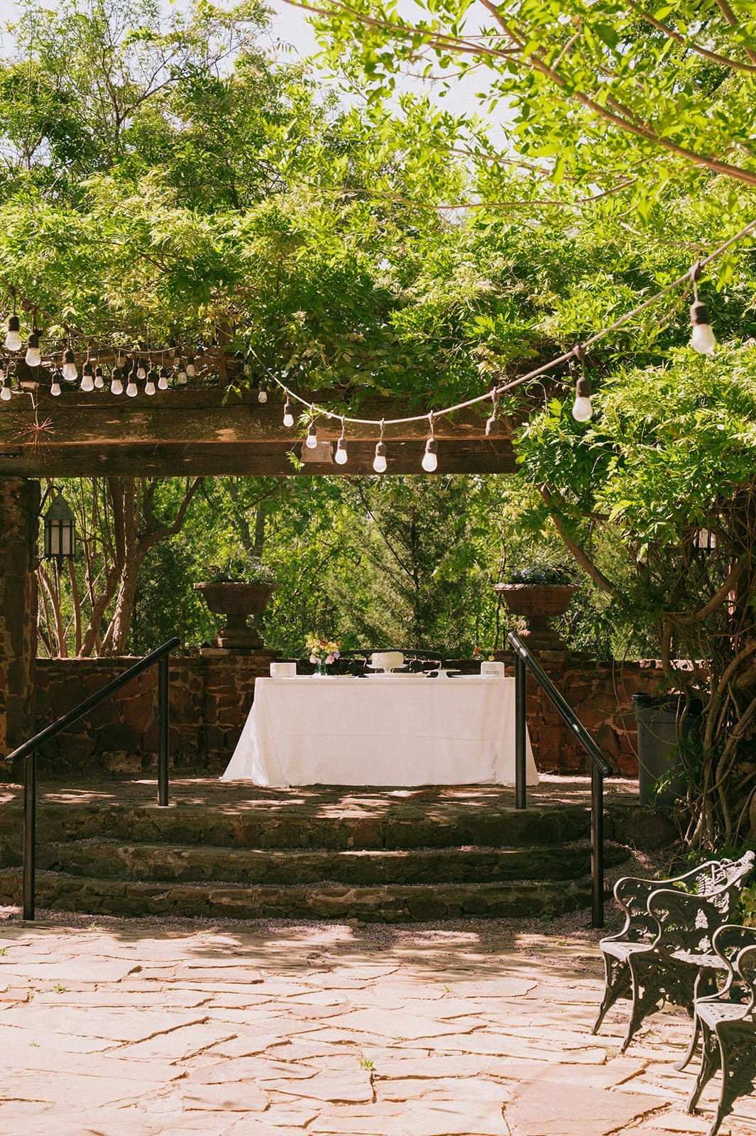 Outdoor patio with stone steps, a white table covered with a cloth, string lights overhead, and green trees surrounding the area.