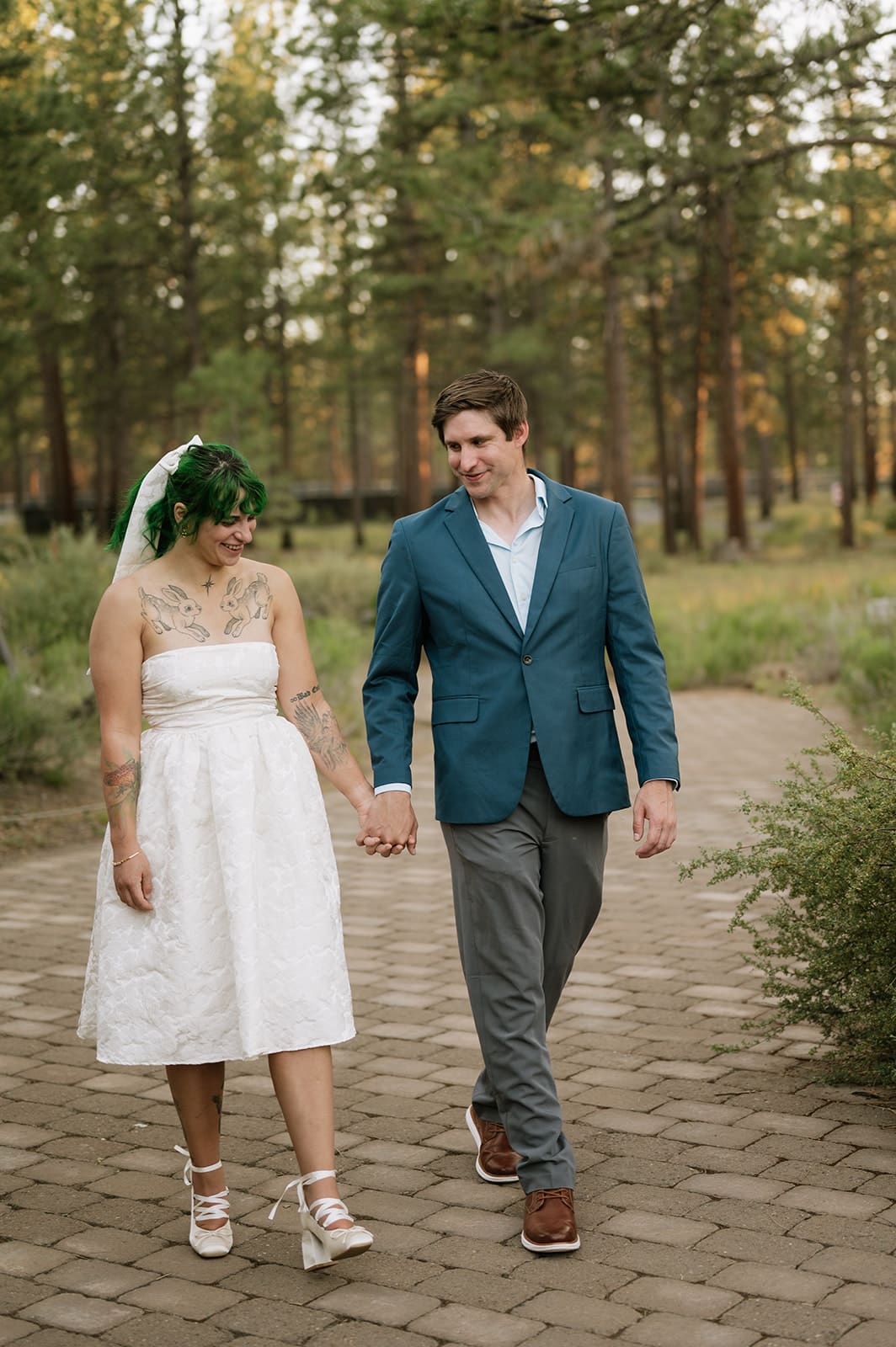 A couple walks hand in hand on a stone path in a wooded area. The woman wears a white dress and veil; the man wears a blue jacket, white shirt, and gray pants.