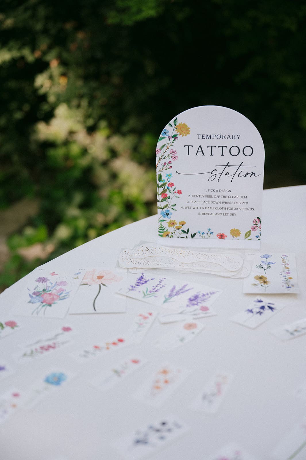 A table displays various floral temporary tattoos and a sign reading “Temporary Tattoo Station” outdoors with greenery in the background.