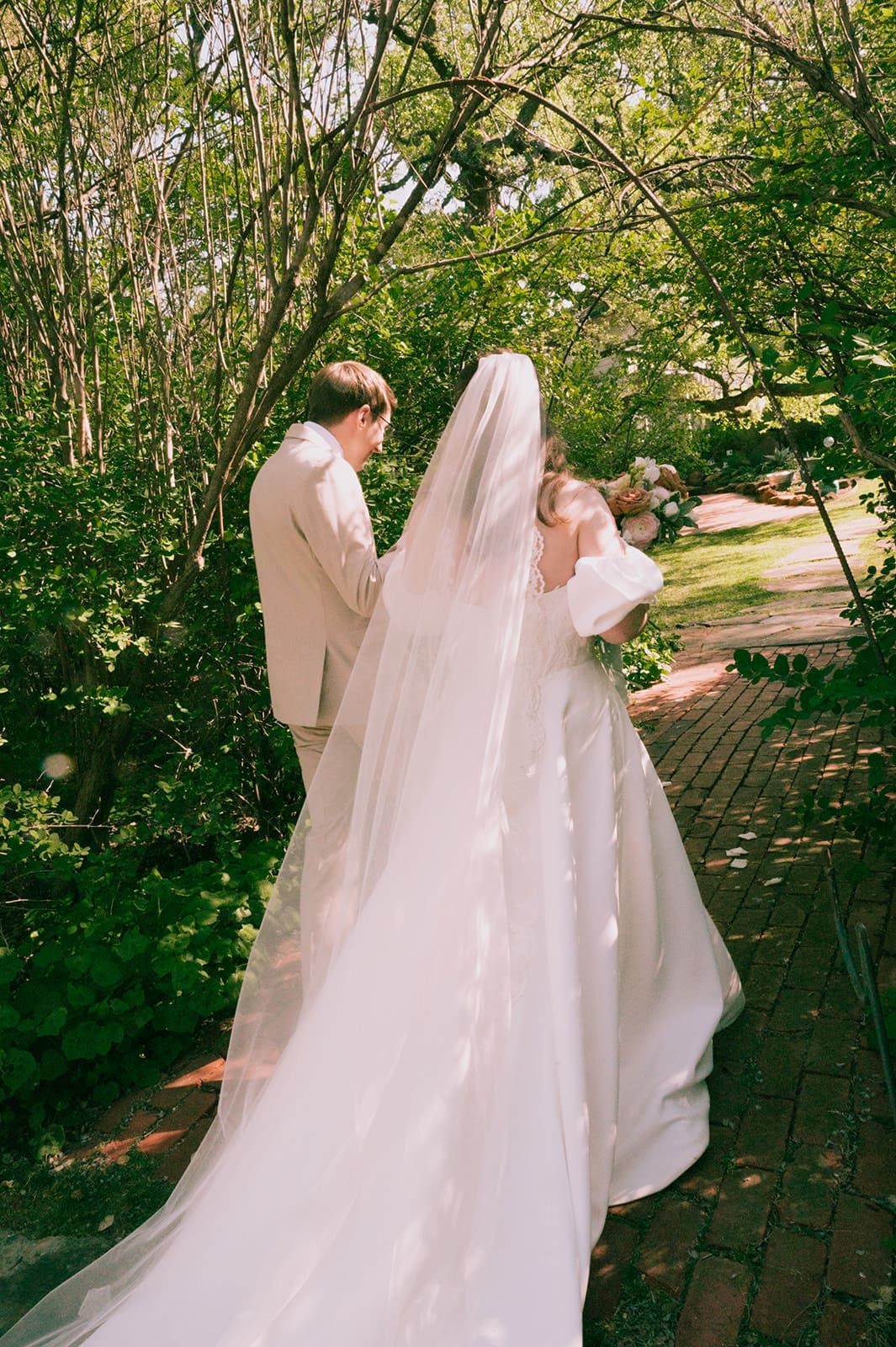 A bride in a white dress holds a bouquet of pastel and brown flowers, standing next to a person in a beige suit.