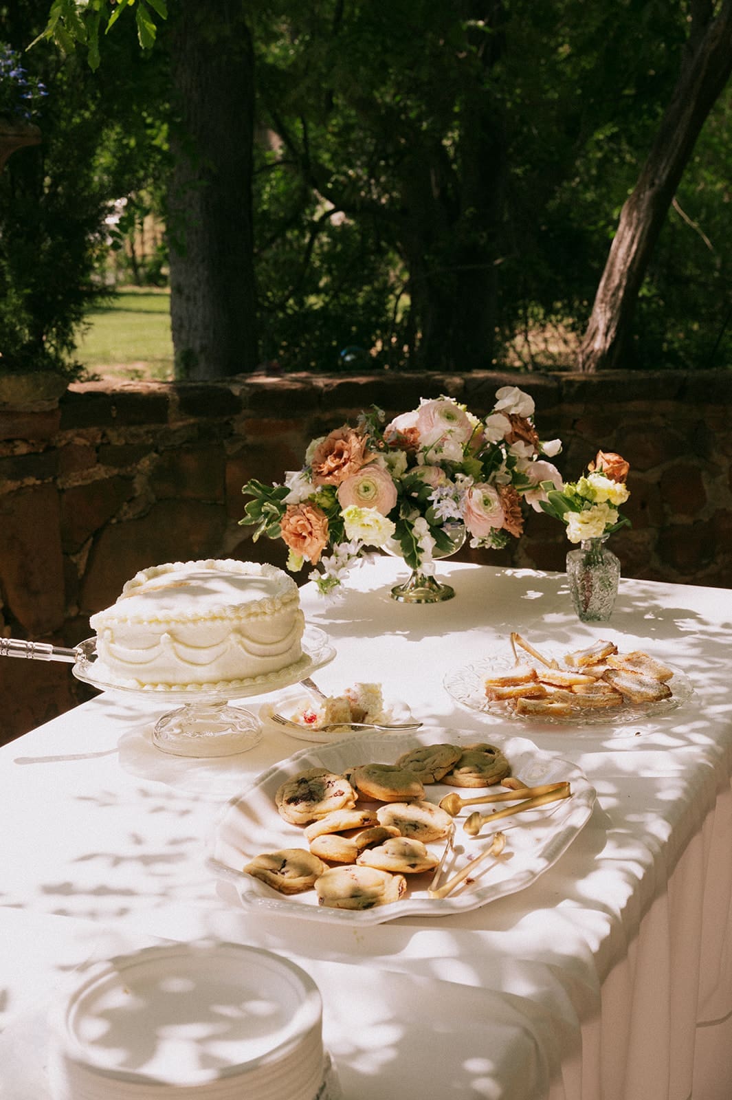 A decorated outdoor table displays a white cake, assorted pastries, and a floral arrangement under tree shade.