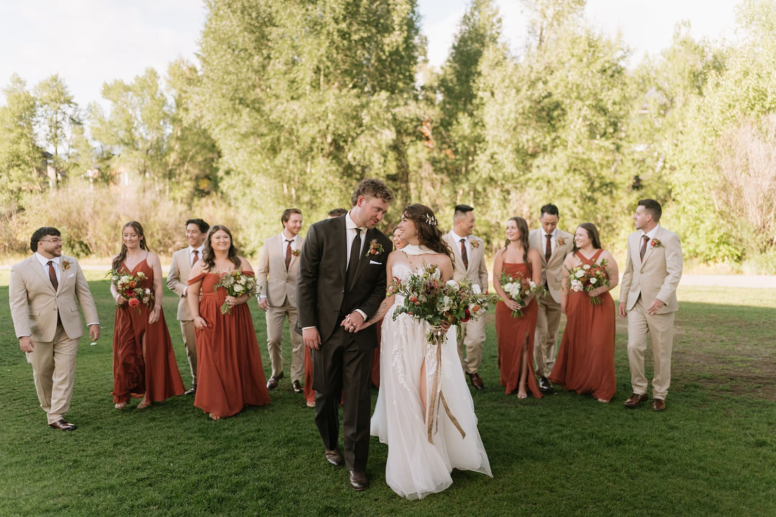 A bride and groom walk hand in hand through a park, followed by their wedding party.