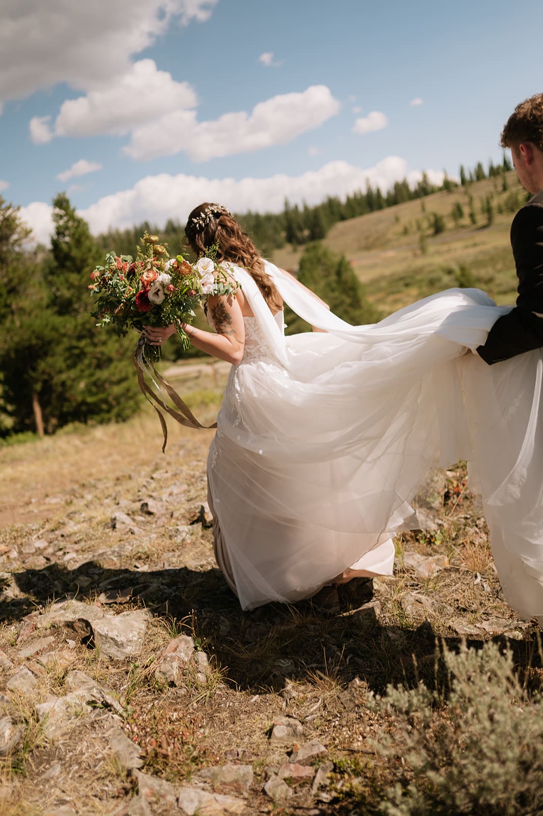 A bride holding a bouquet walks through a grassy field while her partner helps carry the train of her dress.