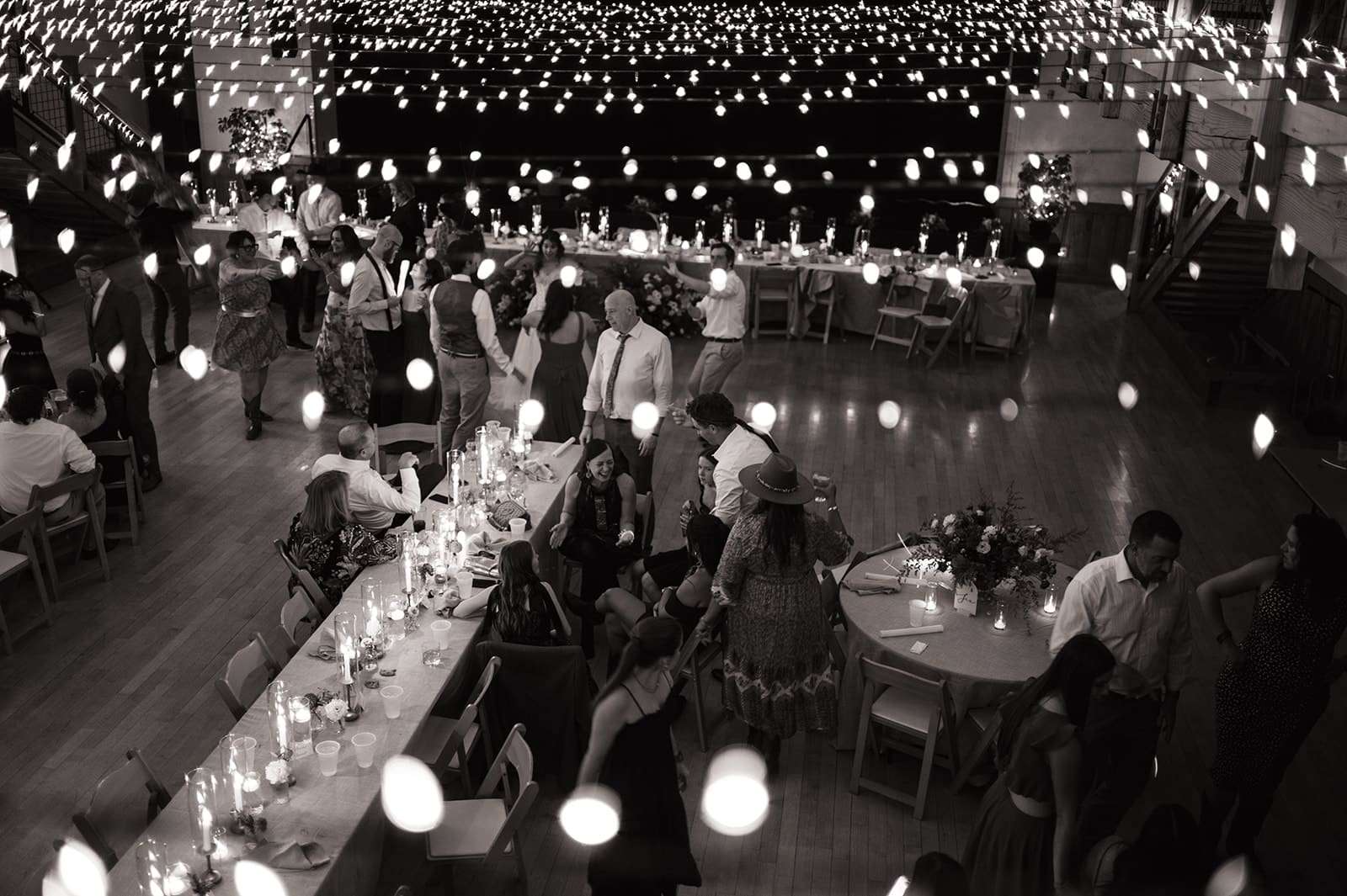 A high-angle black and white shot of guests gathered at long tables in a room decorated with hanging string lights.
